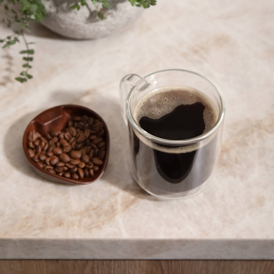 Clear glass mug with coffee and a small bowl of coffee beans on a marble surface.