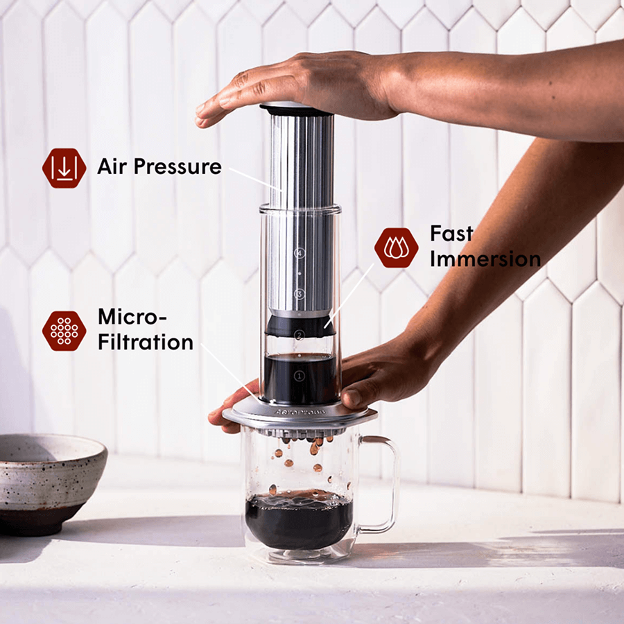 Person using an AeroPress Premium coffee maker with text labels on air pressure, fast immersion, and micro-filtration. #color_silver
