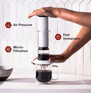 Person using a white AeroPress Premium coffee maker with text labels for air pressure, fast immersion, and micro-filtration. #color_white