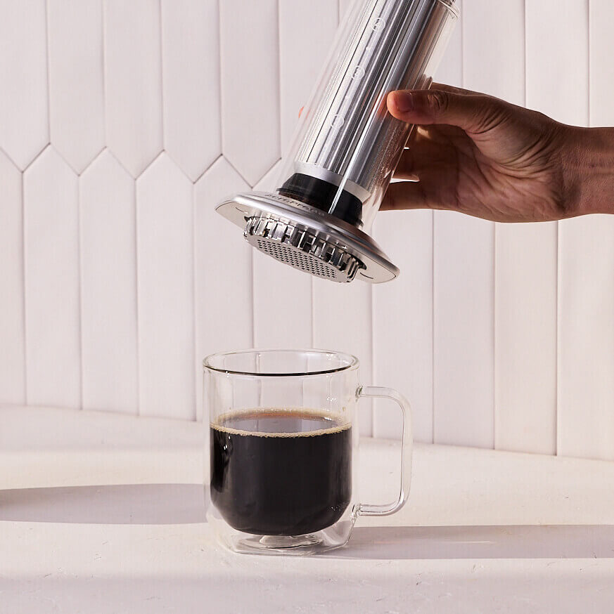 Hand using an AeroPress Premium to brew coffee into a clear mug on a white countertop.