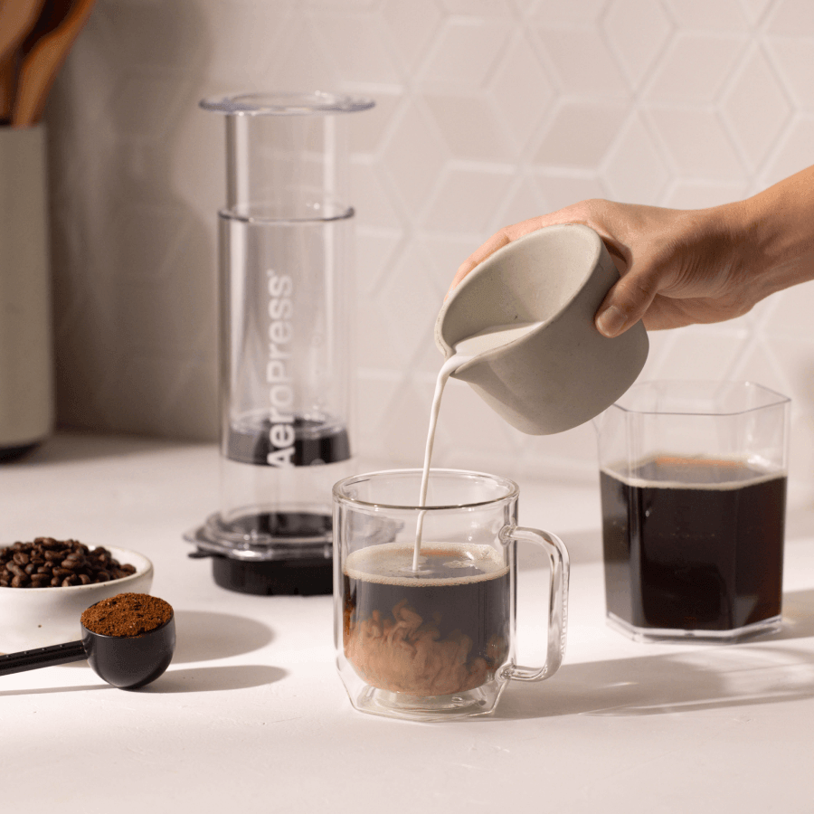 Person pouring milk into a glass of coffee with a AeroPress in the background