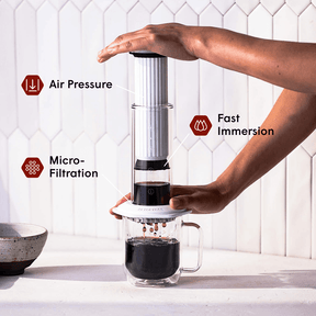 Person using am AeroPress Premium coffee maker with text labels for air pressure, fast immersion, and micro-filtration. #color_white