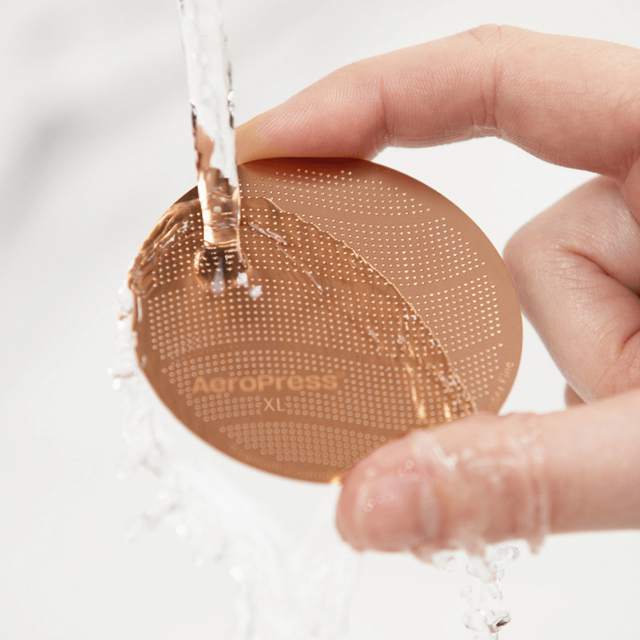 A person holding an AeroPress Gold Tone Filter under running water.