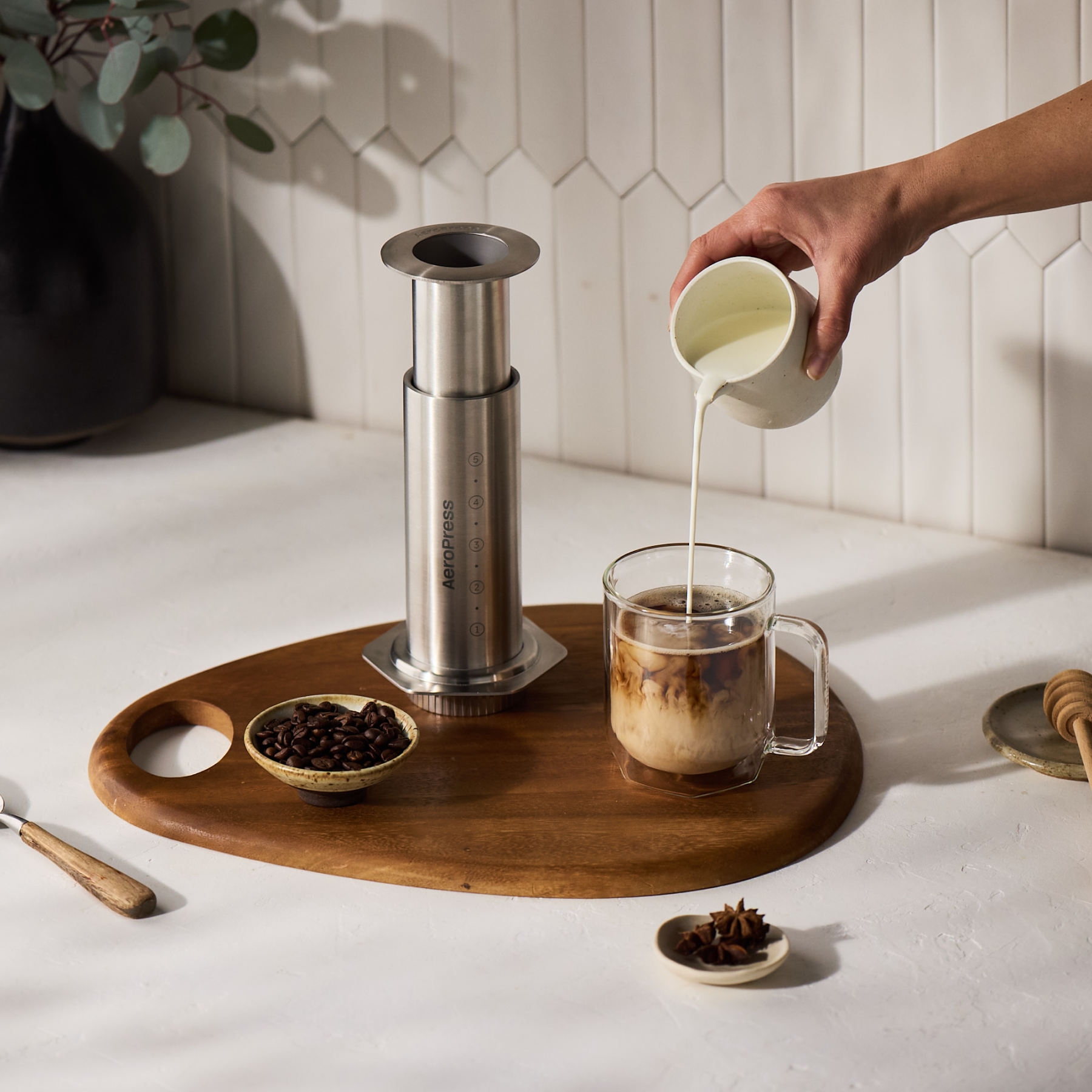 Person pouring milk into a coffee cup on a wooden tray with a AeroPress Steel coffee maker and coffee beans.
