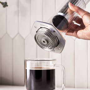 Hand pouring coffee from a clear glass AeroPress Premium coffee press into a glass mug with a white tiled wall background.