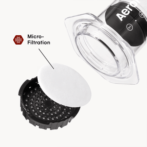 AeroPress Clear coffee maker next to a paper filter and filter cap on a white background