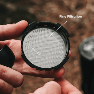Person holding an AeroPress filter cap with a stainless steel filter inside of it with a natural background