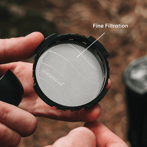 Person holding an AeroPress filter cap with a stainless steel filter inside of it with a natural background