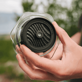 Person placing an AeroPress Flow Control Cap onto an AeroPress Clear chamber with a blurred background of trees