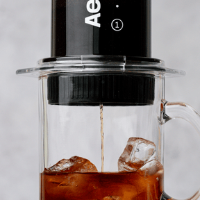 Up close shot of an AeroPress Clear and Flow Control Cap being used to brew coffee directly over ice in a glass mug on a light grey background