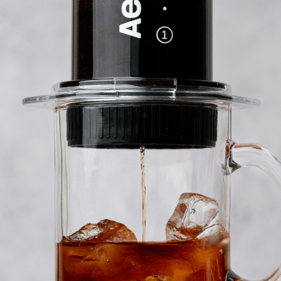 Up close shot of an AeroPress Clear and Flow Control Cap being used to brew coffee directly over ice in a glass mug on a light grey background