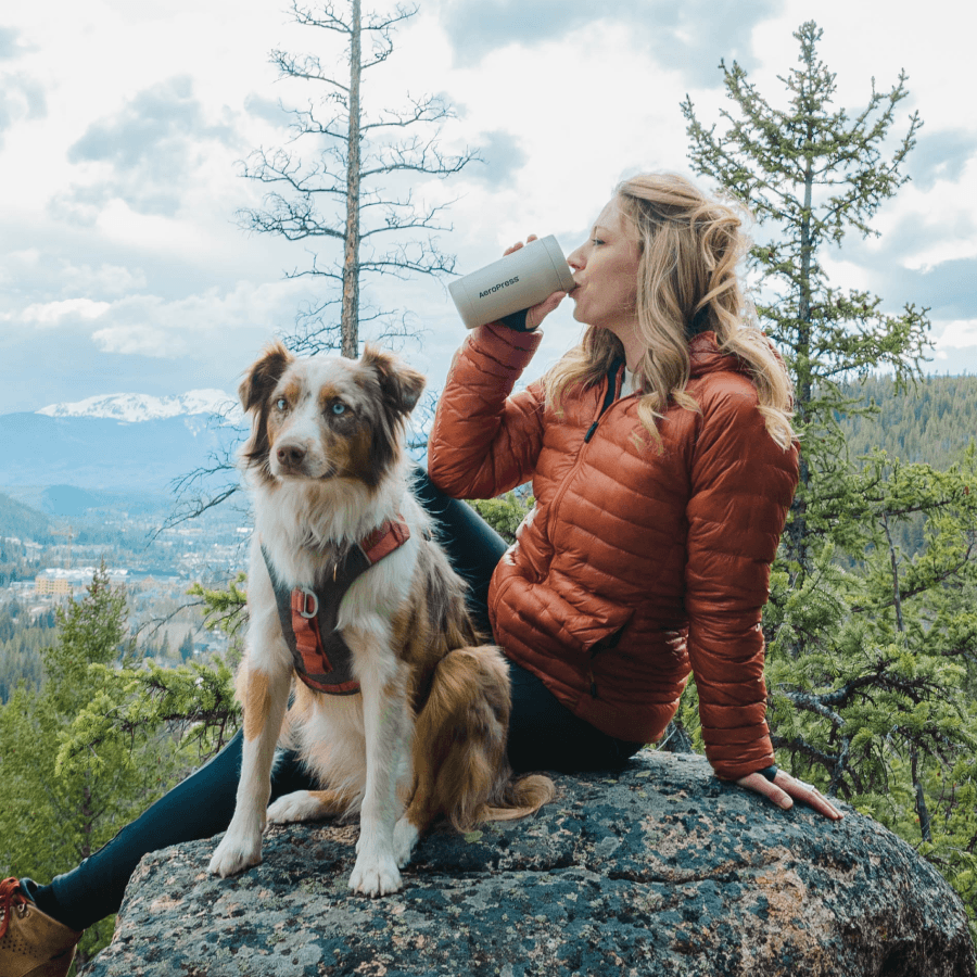 Woman in orange jacket drinking from an AeroPress Tumbler with a dog on a rocky outcrop in a forested area. #color_cream