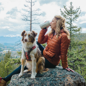 Woman in orange jacket drinking from an AeroPress Tumbler with a dog on a rocky outcrop in a forested area. #color_black