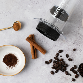 AeroPress coffee maker with coffee beans, ground coffee, and cinnamon sticks on a light gray surface. #color_green