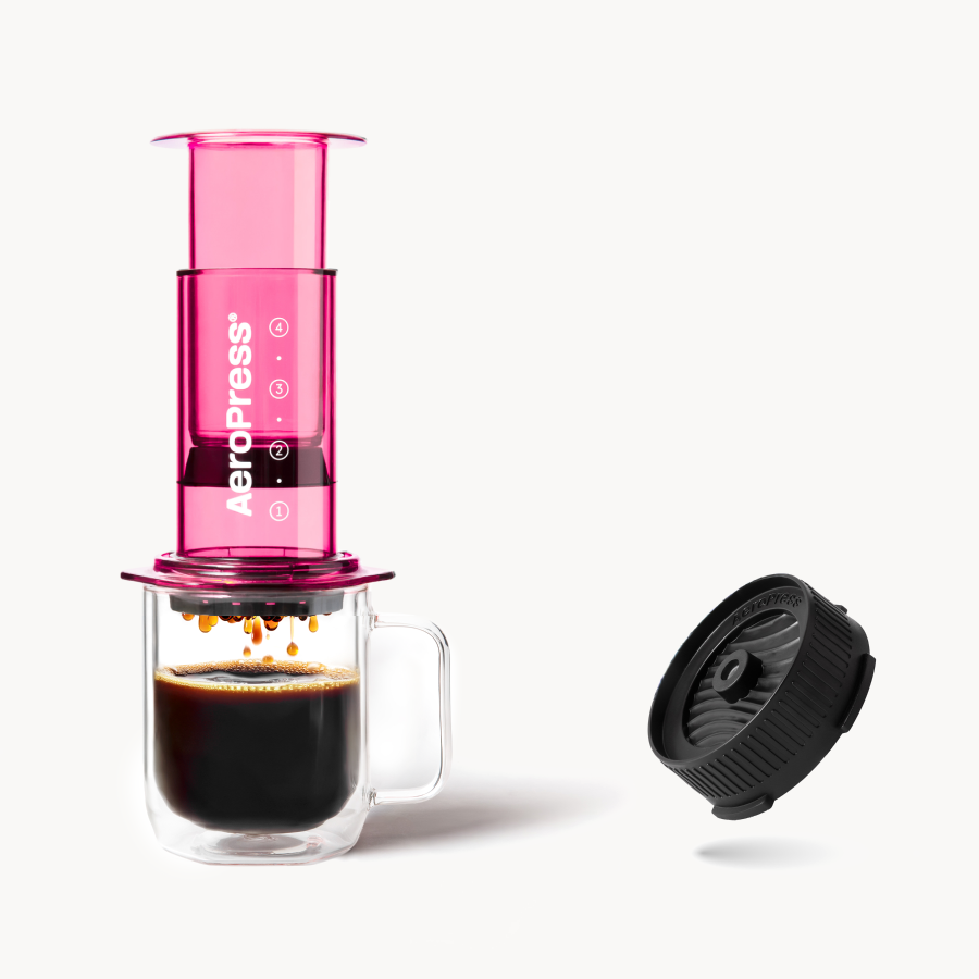 Pink AeroPress coffee maker with a glass mug of coffee and a Flow Control Cap on a white background #color_pink