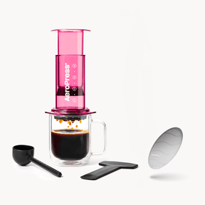 Pink AeroPress coffee maker with a glass mug, scoop, and filter on a white background #color_pink
