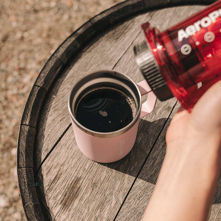 Person pouring coffee from a red Aeropress into a pink mug on a wooden surface. #color_pink