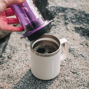 Person pouring coffee from a purple AeroPress into a white mug on a concrete surface.  #color_purple