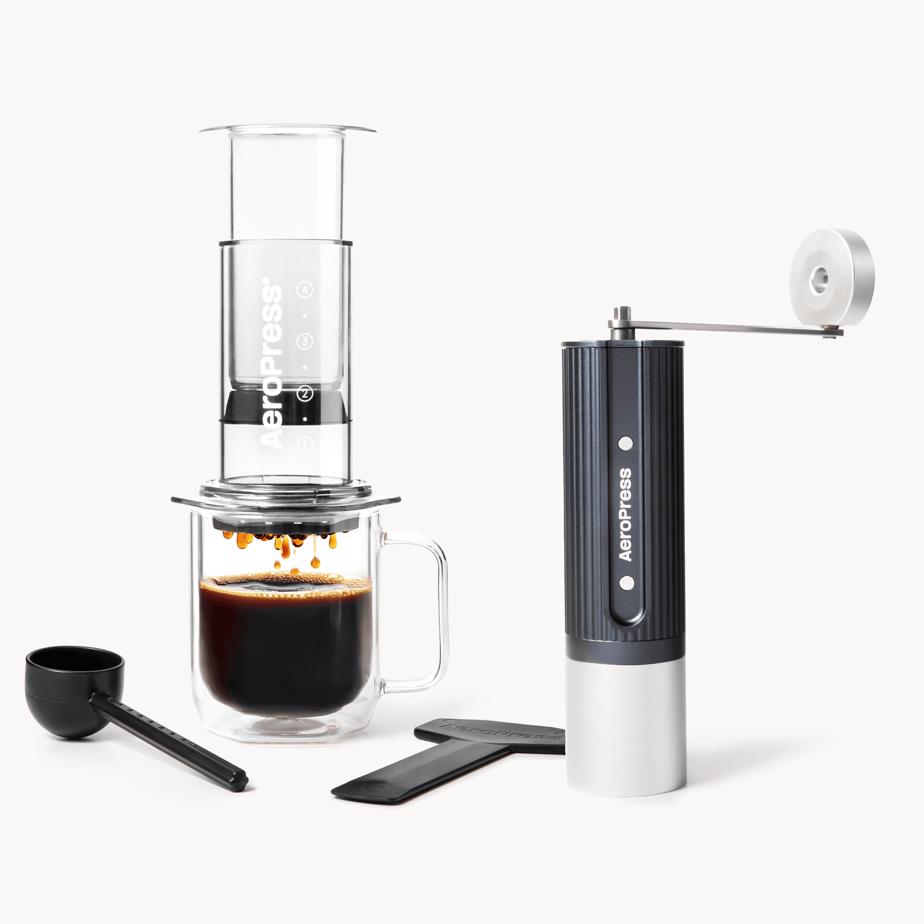 Aeropress coffee maker with a glass of brewed coffee and a handheld grinder on a white background #color_Clear