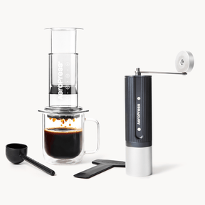 Aeropress coffee maker with a glass of brewed coffee and a handheld grinder on a white background #color_Clear