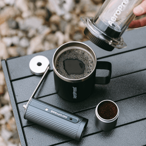 Aeropress coffee maker being used to pour coffee into a black mug on a wooden surface. #color_Clear