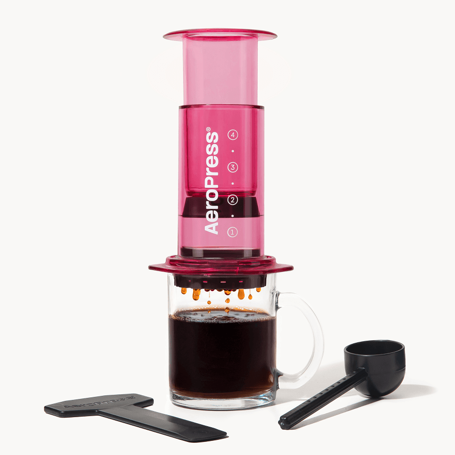 Pink AeroPress coffee maker with a glass mug of coffee and scoop on a white background Clear Pink #color_pink