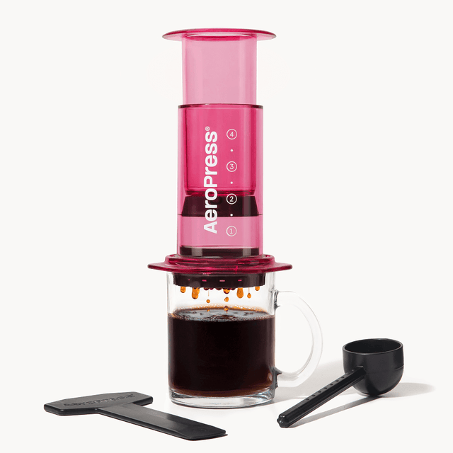 Pink AeroPress coffee maker with a glass mug of coffee and scoop on a white background Clear Pink #color_pink
