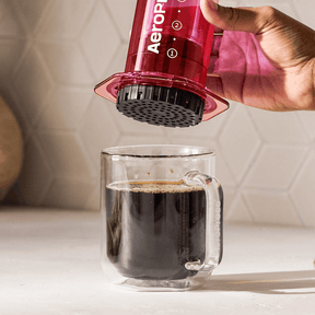 Person using an Aeropress to make coffee in a clear mug. #color_pink