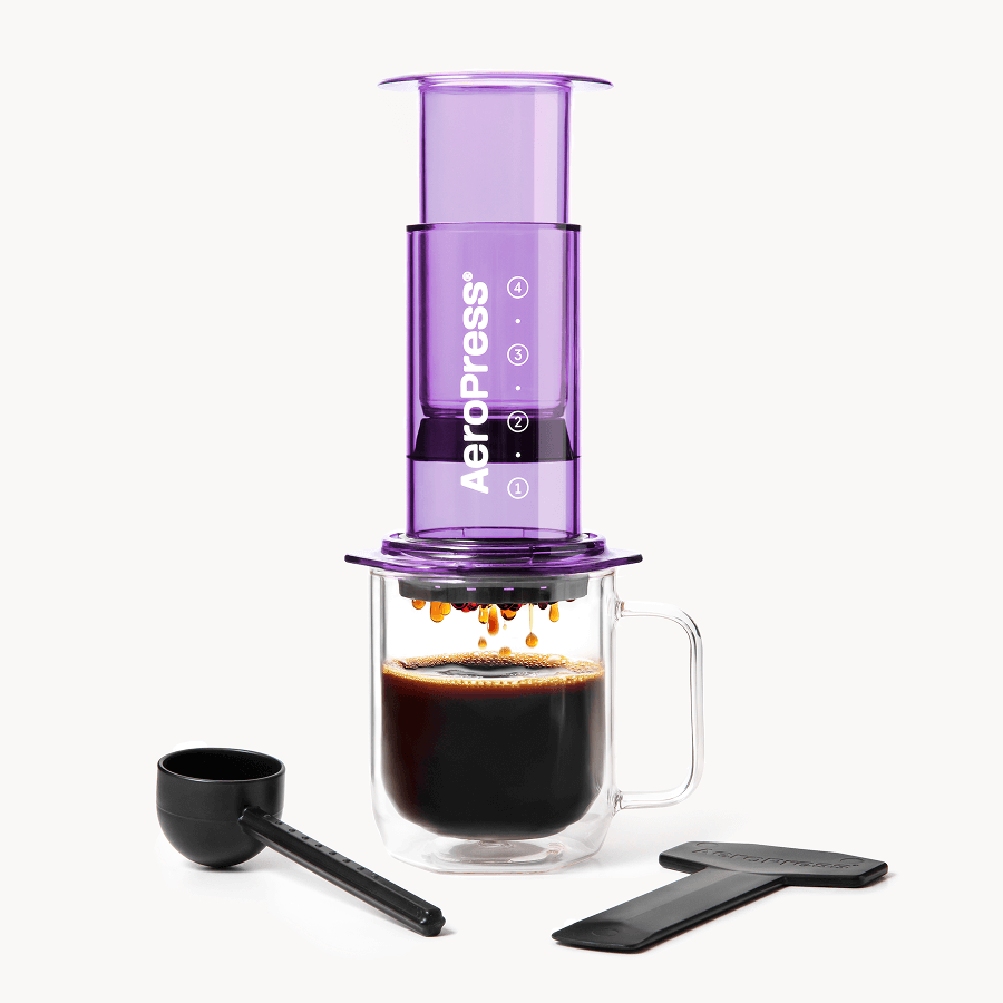 Purple AeroPress coffee maker with a glass mug of coffee and scoop on a white background #color_purple