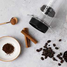 AeroPress coffee maker with coffee beans, ground coffee, and cinnamon sticks on a light gray surface. #color_clear