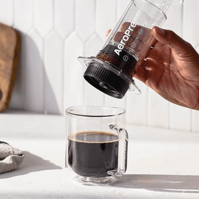 Person pouring coffee from an Aeropress into a glass mug on a light wooden surface.#color_clear