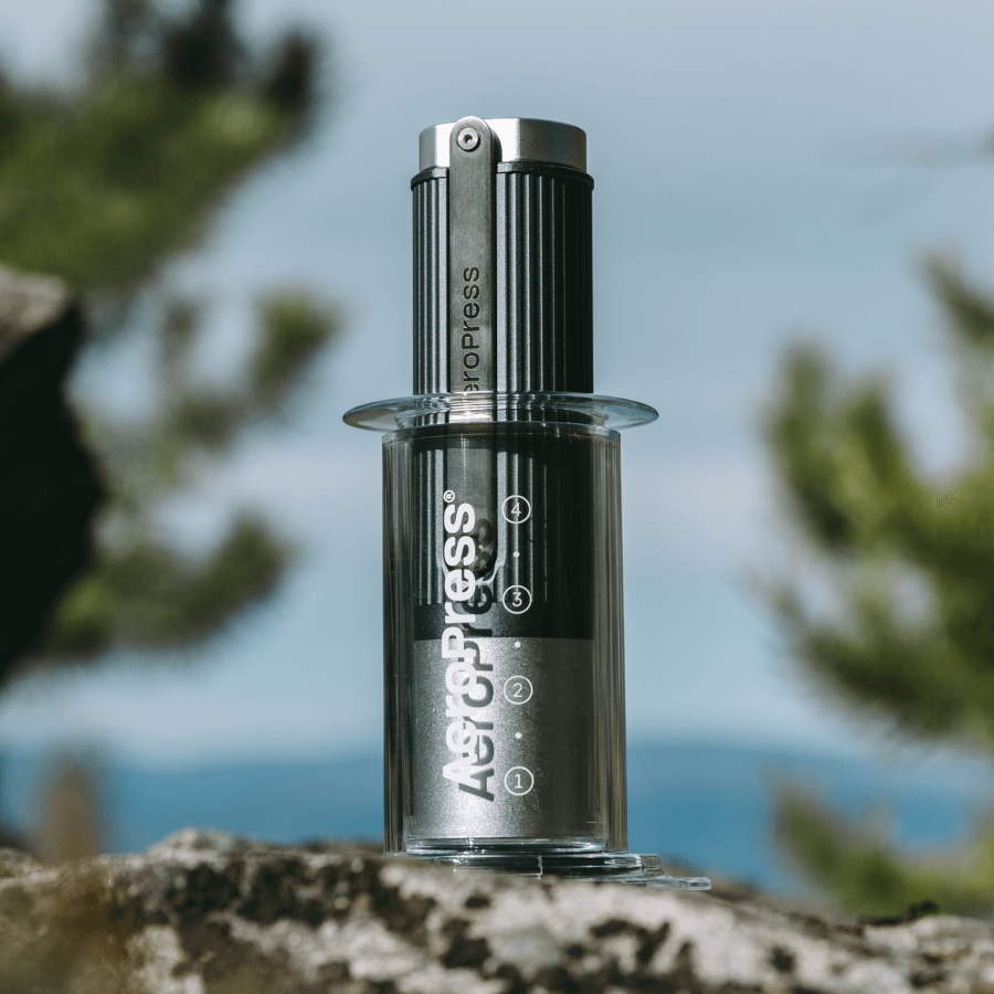 AeroPress Clear coffee maker with the AeroPress manual grinder inside of it on a rock with a blurred natural background