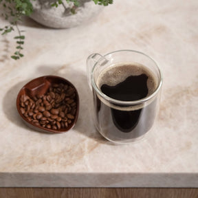 Clear glass mug with coffee and a small bowl of coffee beans on a marble surface.