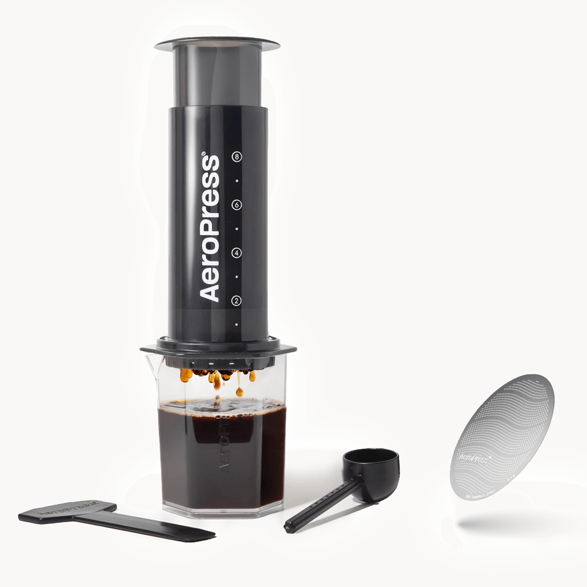 Aeropress coffee maker with a cup of coffee, scoop, and filter on a white background