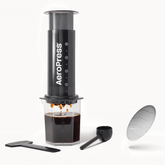 Aeropress coffee maker with a cup of coffee, scoop, and filter on a white background