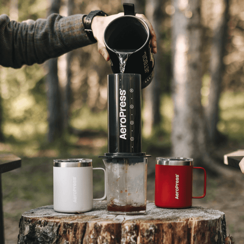 Person pouring coffee from an Aeropress into a mug outdoors.