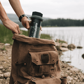 Person removing an AeroPress Original XL coffee maker from a backpack next to a lake.