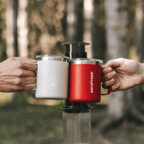 Two people clinking AeroPress stainless steel mugs together in a cheers against a forest background with an AeroPress Original XL behind them.