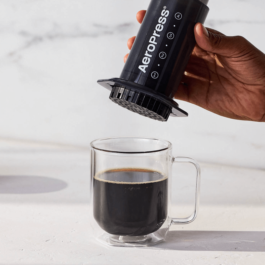 A person using an AeroPress Original to pour coffee into a clear glass mug on a light background