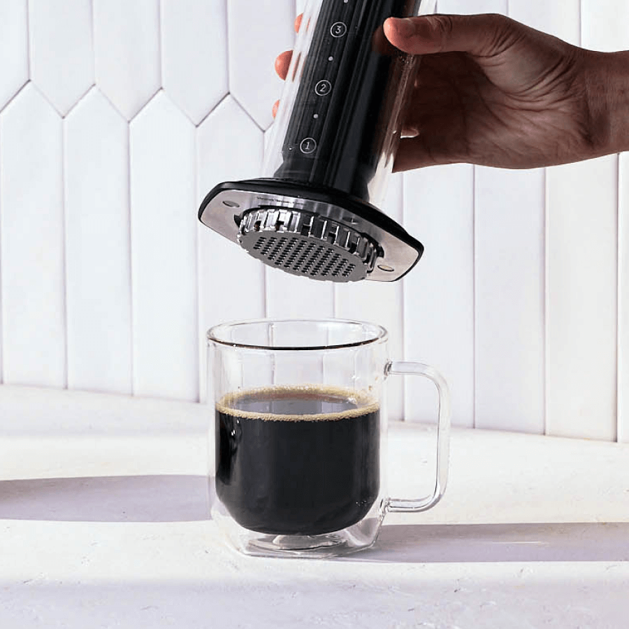 Hand using a AeroPress Premium coffee maker to pour coffee into a glass mug against a white tiled wall. #color_black