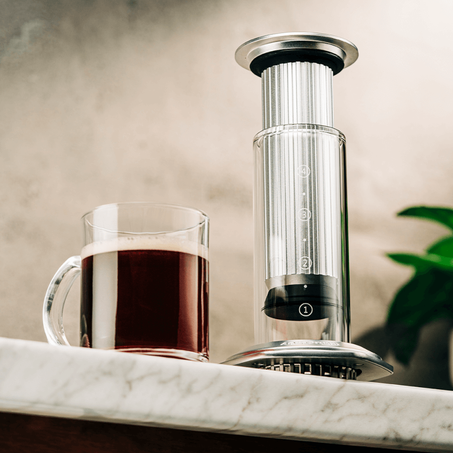 Silver AeroPress Premium coffee maker with a glass of coffee on a marble surface