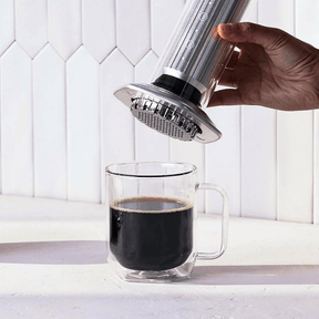 Hand using a AeroPress Premium to pour coffee into a clear glass mug on a white background. #color_silver