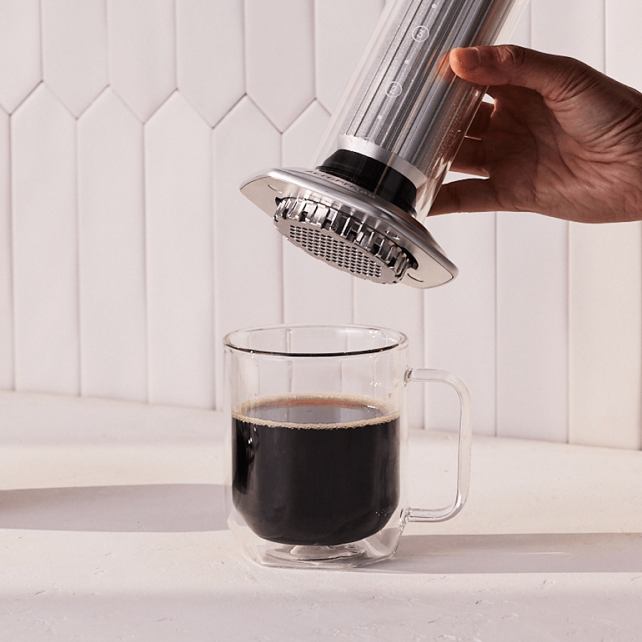 Hand using a AeroPress Premium to pour coffee into a clear glass mug on a white background. #color_silver