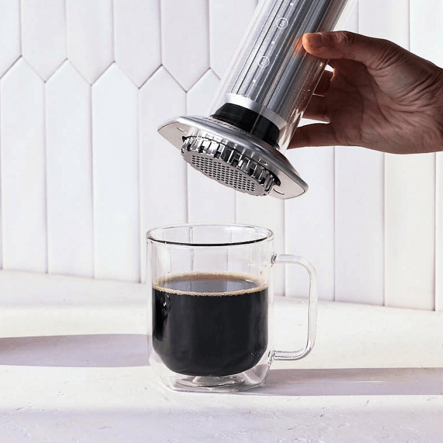 Hand using a AeroPress Premium to pour coffee into a clear glass mug on a white background. #color_silver