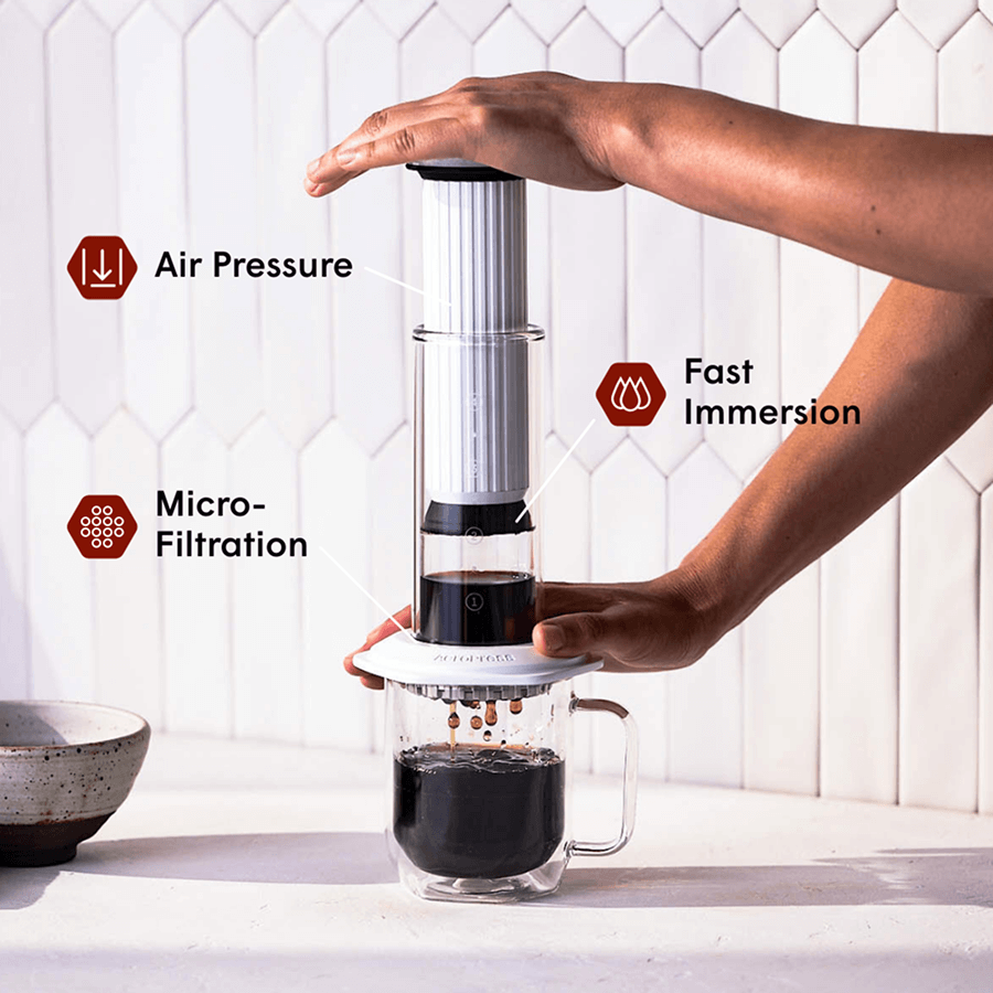 Person using a white AeroPress Premium coffee maker with text labels for air pressure, fast immersion, and micro-filtration. #color_white