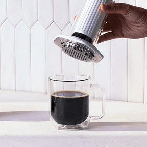 Hand using an AeroPress Premium coffee maker to pour coffee into a clear glass mug on a white countertop. #color_white