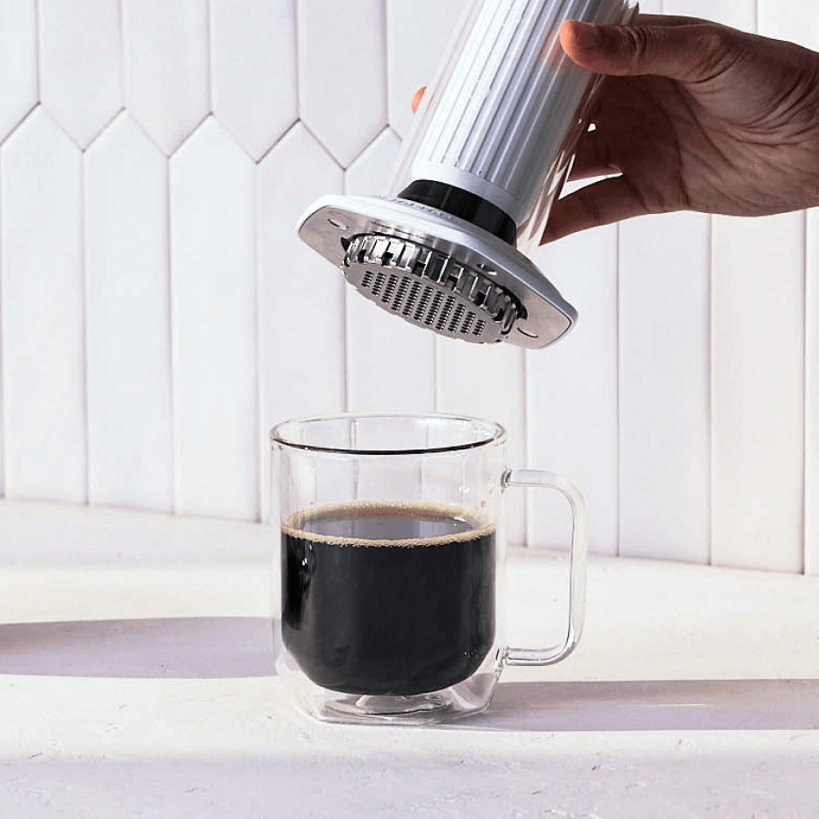 Hand using an AeroPress Premium coffee maker to pour coffee into a clear glass mug on a white countertop. #color_white