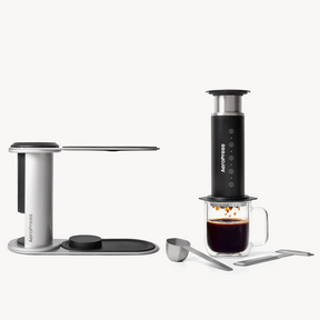 AeroPress coffee maker with a glass of coffee and accessories on a white background  #color_black
