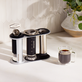Aeropress coffee maker with three pods and a glass of coffee on a light  surface. #color_black
