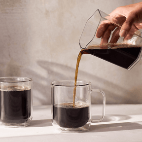 Person pouring coffee from a glass carafe into a clear mug on a light surface.
